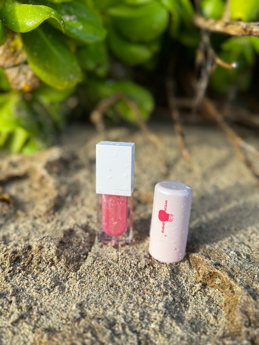 Small pink lip gloss with white cap on sandy surface with green foliage in background. Small light pink makeup product with sugar ocean printed on it.