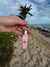 Translation missing: en.Hand holding a pink lip gloss keychain on a beach with palm trees and ocean in the background