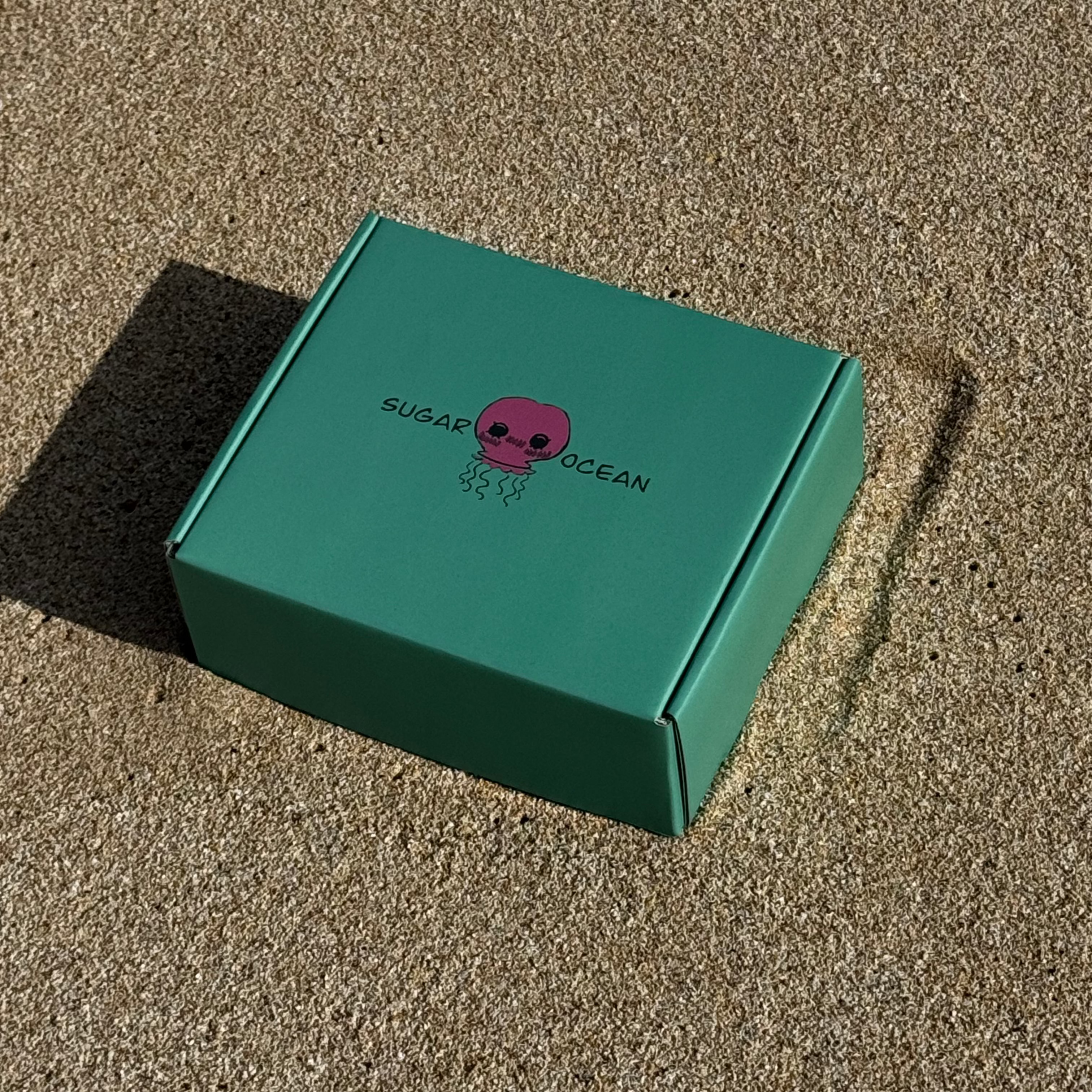 The Current teal subscription box in sand with Sugar Ocean brand name and hot pink jellyfish logo