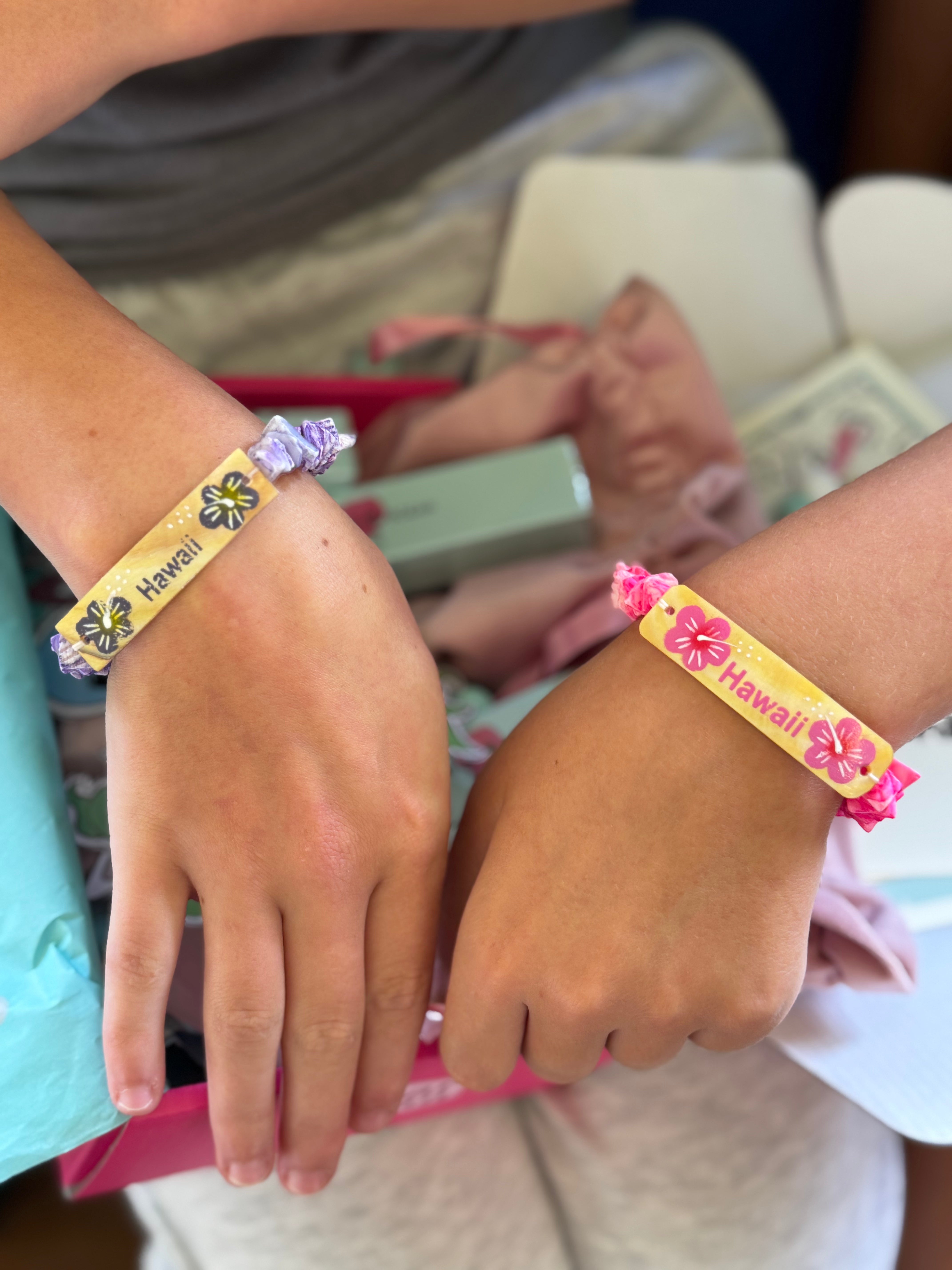 Two hands wearing colorful 'Hawaii' wristbands with a blurred background. friendship gift for the 'twin tides' makeup box.